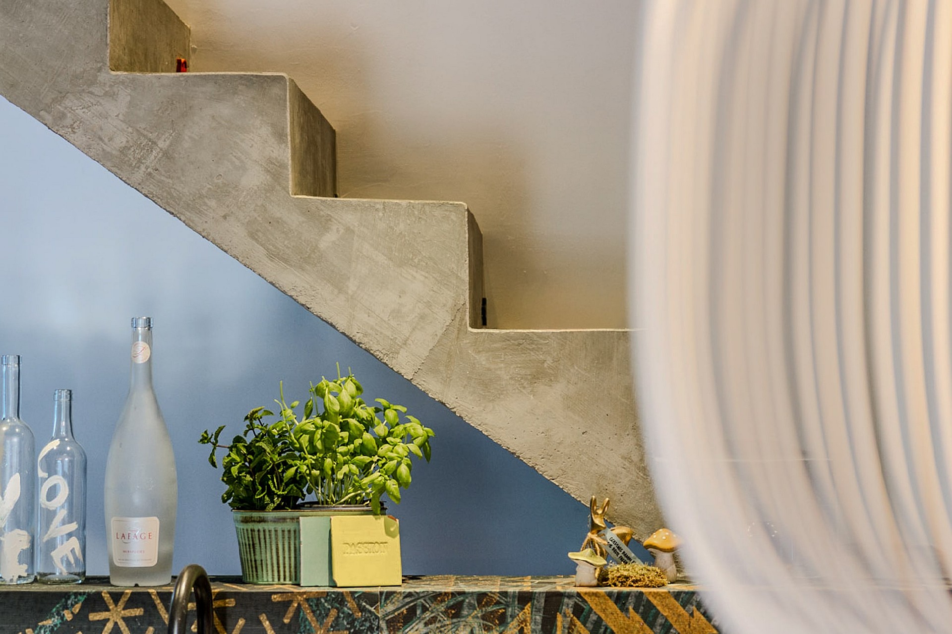 Close-up of a raw concrete staircase with a light blue painted wall, green plant, and glass bottles on a decorative tile ledge.