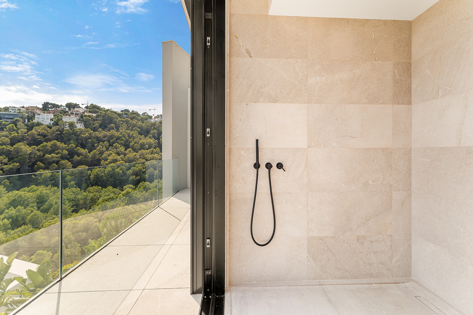 A modern bathroom featuring a walk-in shower with matte black fixtures and a glass-paneled terrace overlooking a green hillside.