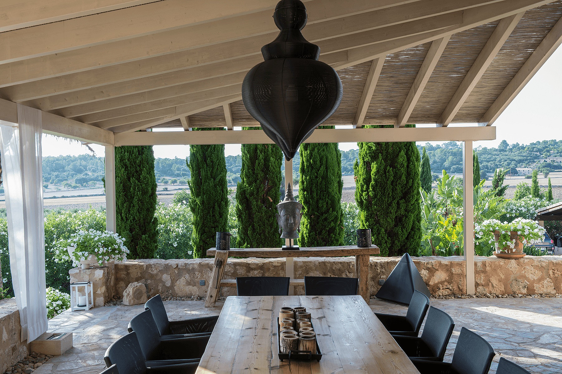 A covered terrace with a long wooden table and chairs, featuring a stone wall, cypress trees, and mountain views in Algaida.