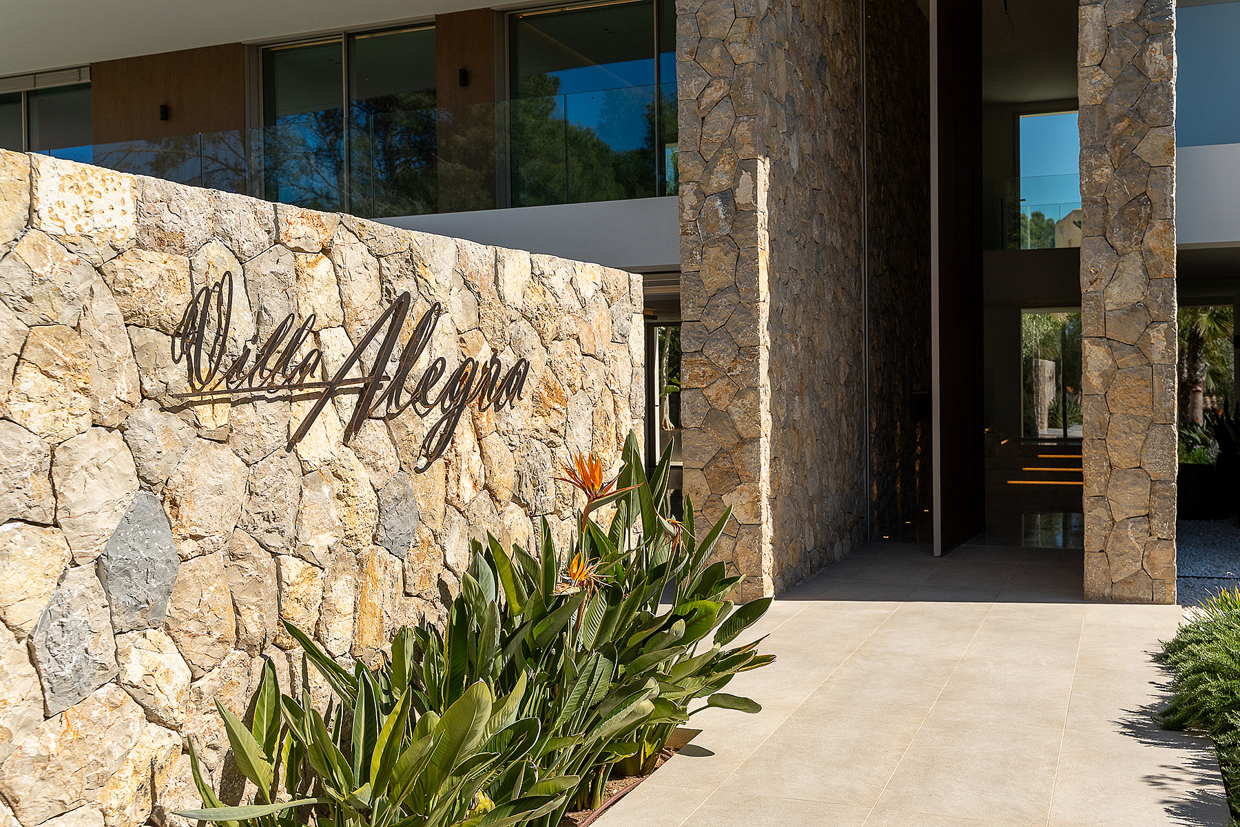 Entrance of a modern villa featuring a dry-stone wall with the name Villa Allegra, stone path, and lush tropical plants.