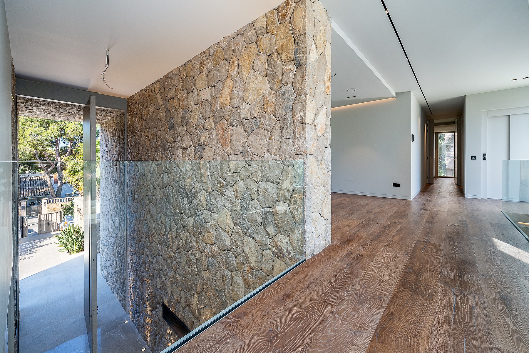 A modern interior hallway features a floor-to-ceiling dry-stone wall, wide oak plank flooring, and a clear glass railing.