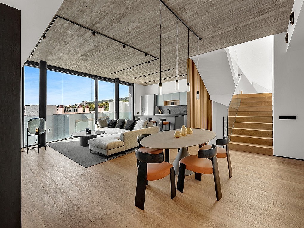 Modern open-plan living and dining room with concrete ceiling, oak flooring, light wood staircase, and floor-to-ceiling windows.