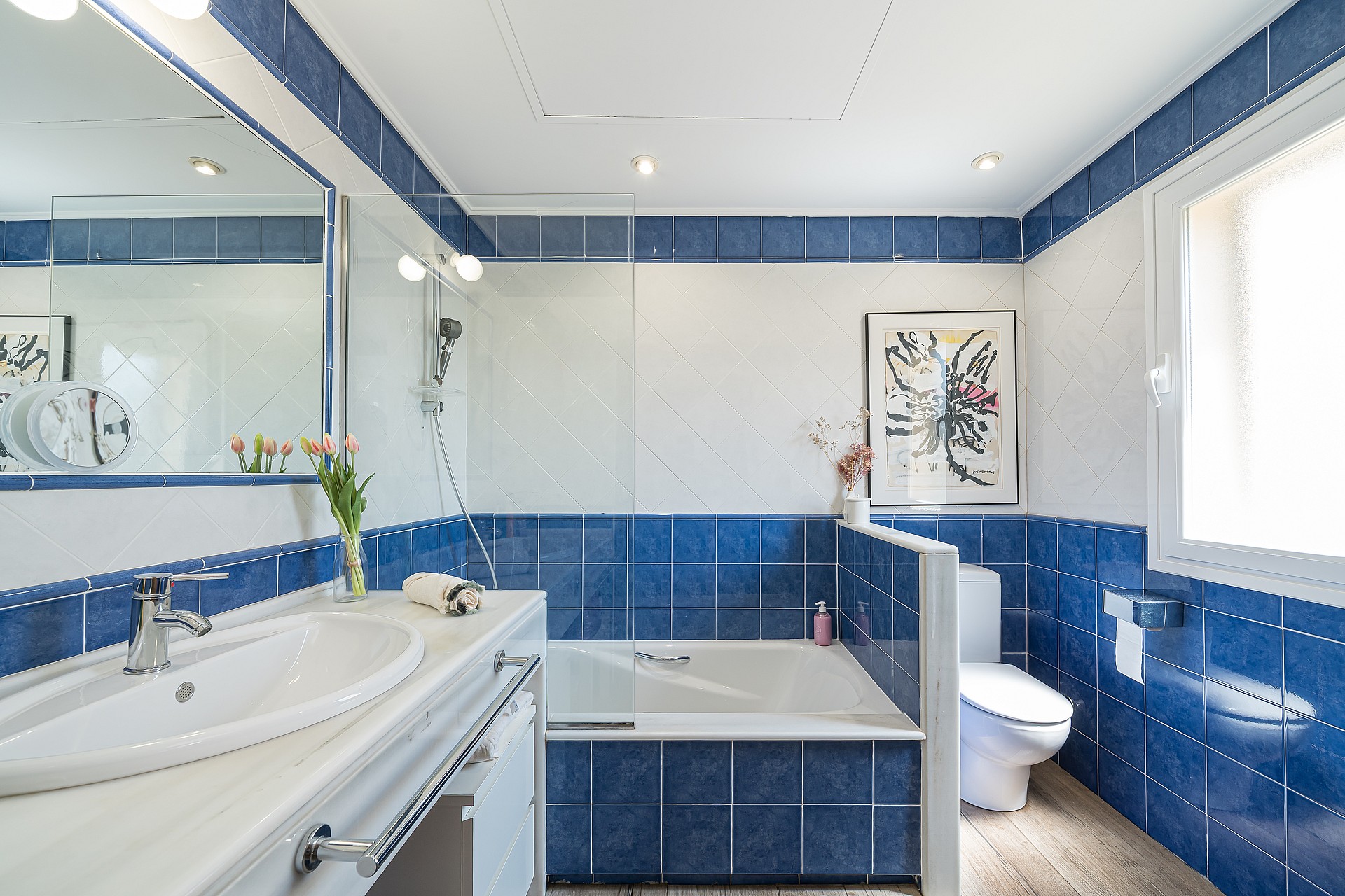 Bright bathroom featuring glossy blue and white wall tiles, a white vanity sink, a built-in bathtub, and a window.
