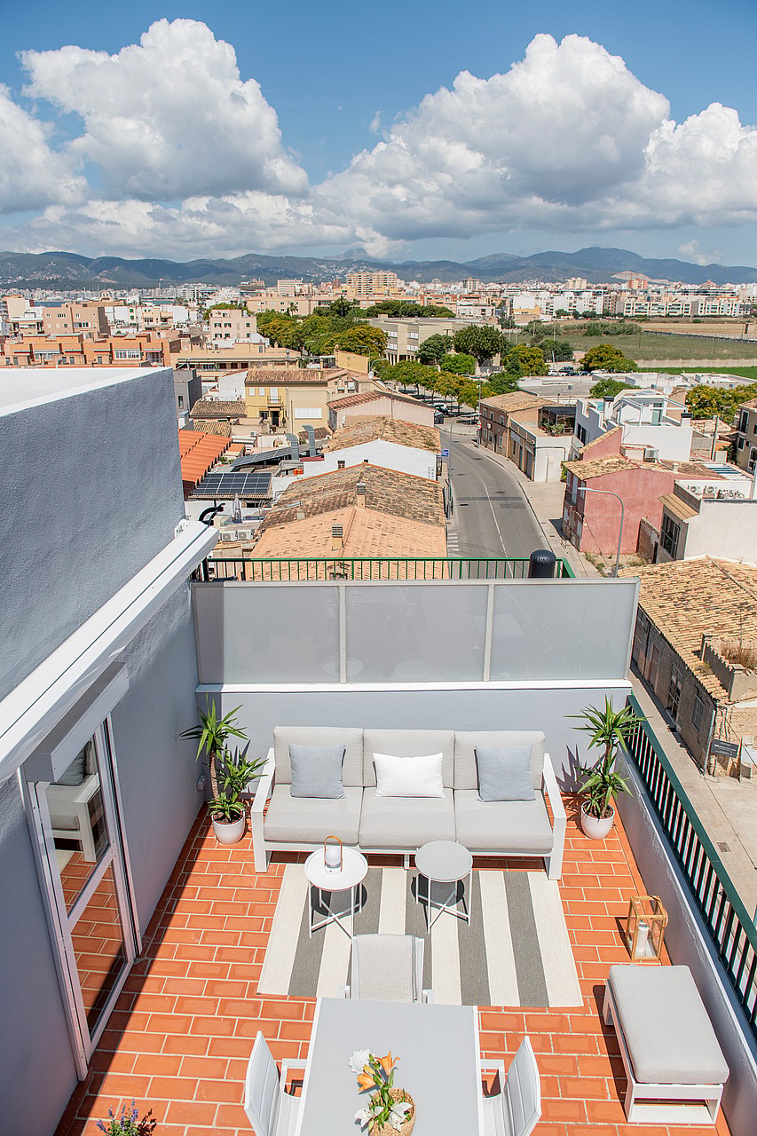 High-angle view of a rooftop terrace with a beige sofa, dining area, and potted plants, overlooking a town and mountains.