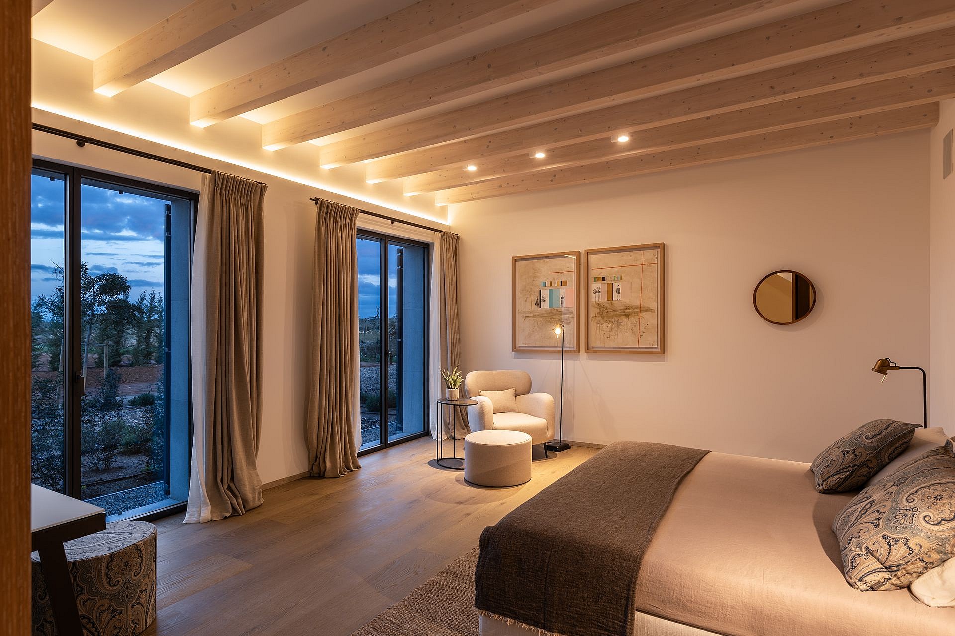 A spacious bedroom with a king-sized bed, exposed wooden ceiling beams, and tall windows showing an evening landscape.
