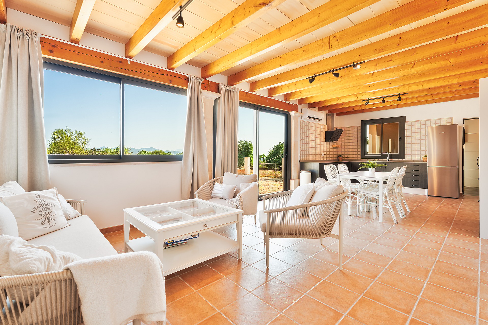 Salón y comedor abierto con vigas de madera vistas, suelos de baldosas y grandes ventanales.