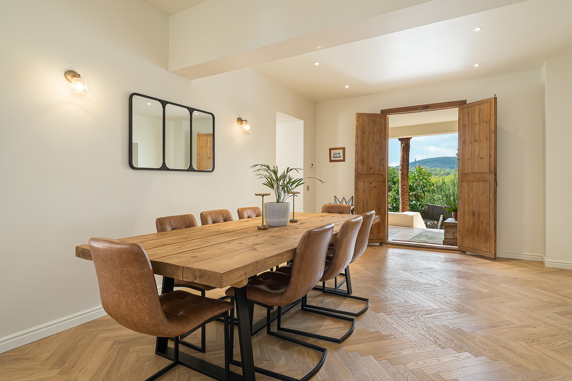 Dining room featuring a long rustic wood table, leather chairs, herringbone flooring, and doors opening to a terrace.
