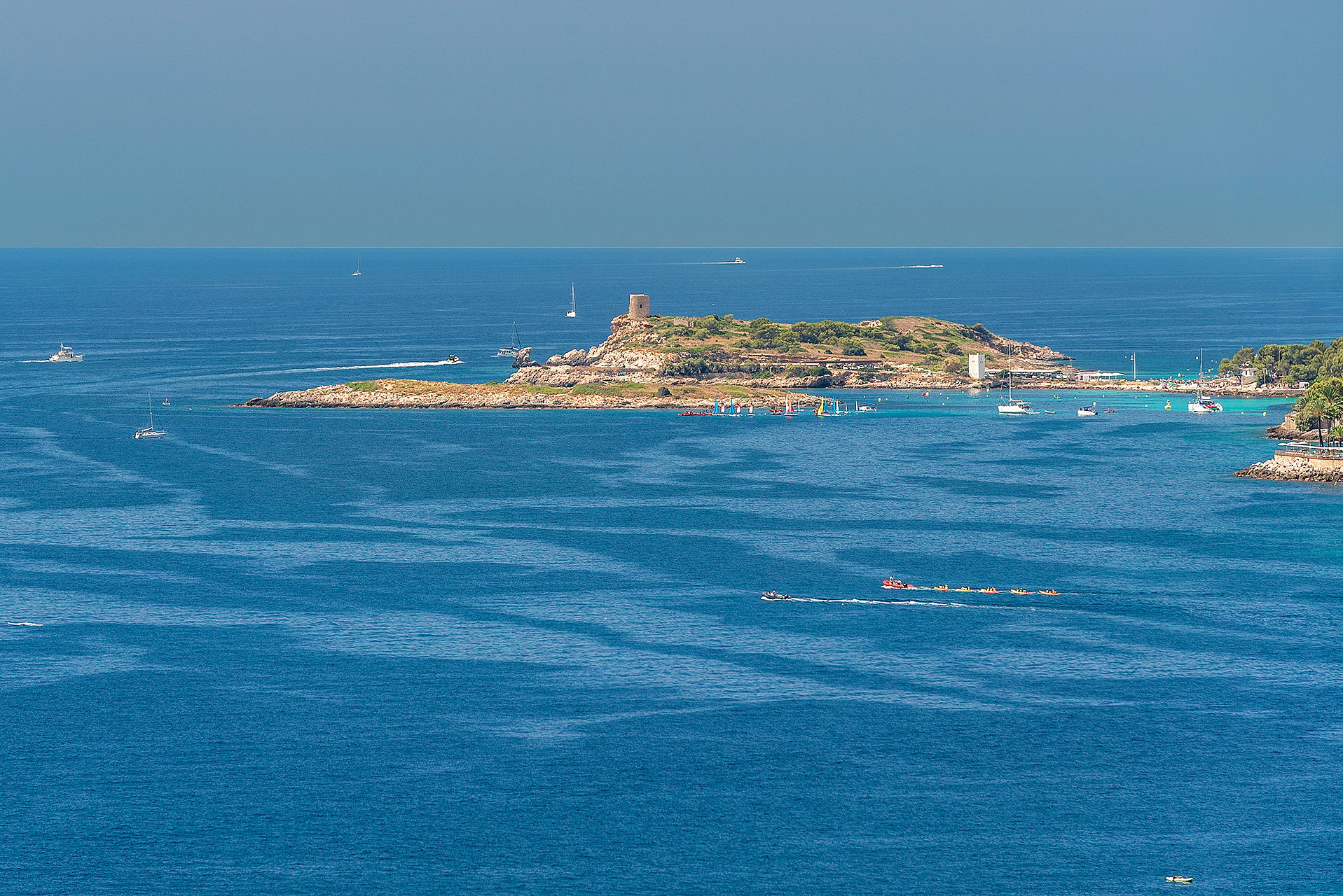 Aerial perspective of the deep blue Mediterranean Sea with a rocky island and distant boats under a clear sky.