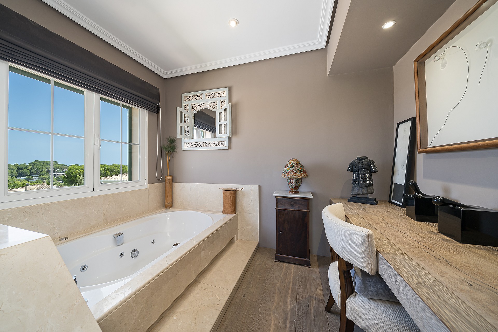 A bright bathroom with a built-in jetted white tub surrounded by beige stone, a stone-topped vanity, and a window with garden views.