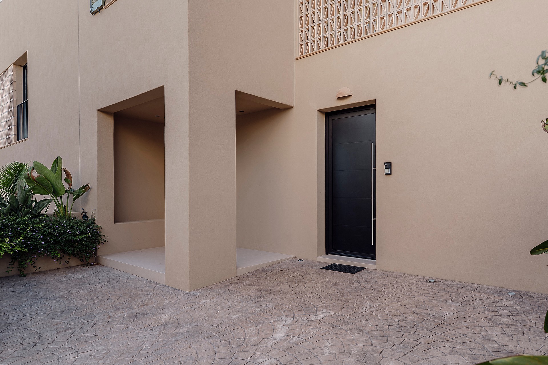 Entrance of a beige townhouse featuring a dark metal front door, gravel pathway, and a decorative concrete block screen.