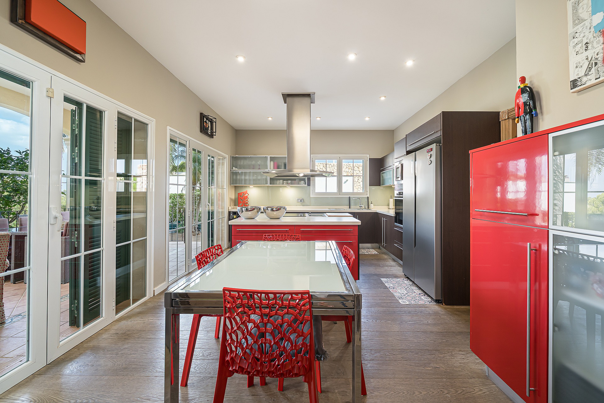 Modern kitchen with red cabinetry, a central dining table, and large glass doors leading to an outdoor terrace.