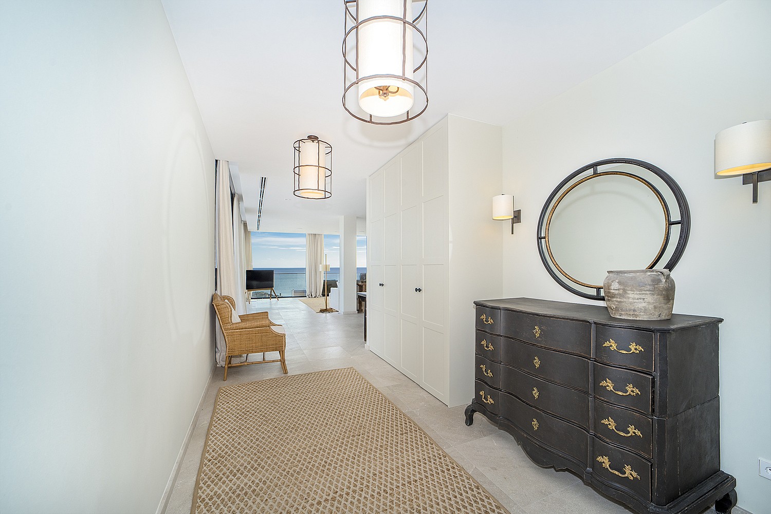 A bright corridor space featuring a dark wood dresser, round mirror, woven rug, and hanging pendant lights.
