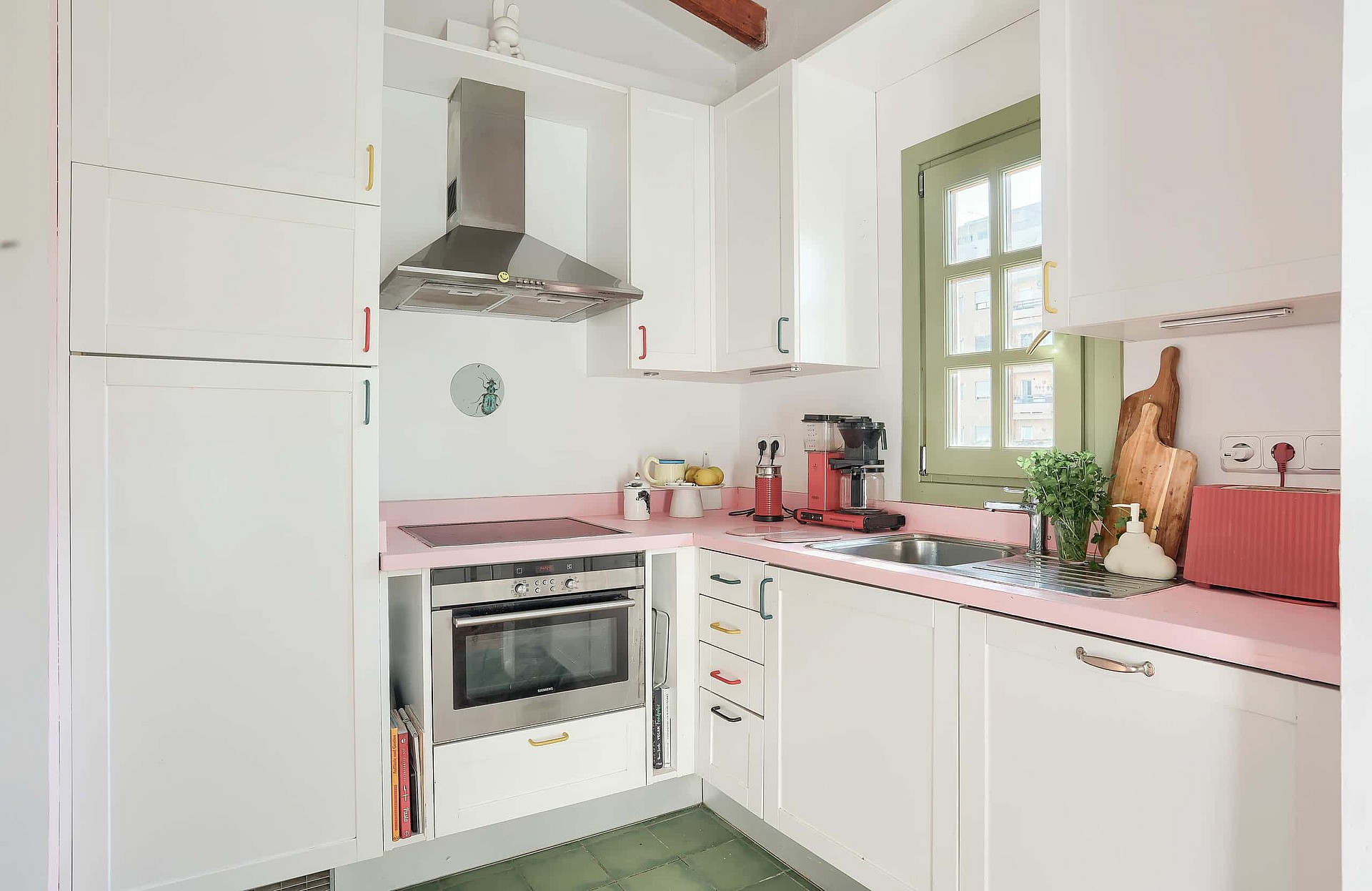 A small white galley kitchen with pale pink countertops, stainless steel appliances, and a green-framed window.