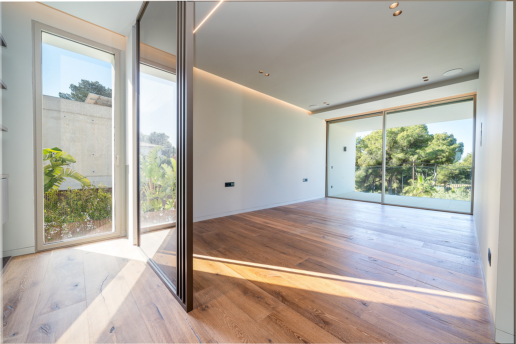 Empty modern room with wide oak floorboards, recessed linear lighting, and large sliding glass doors opening to greenery.
