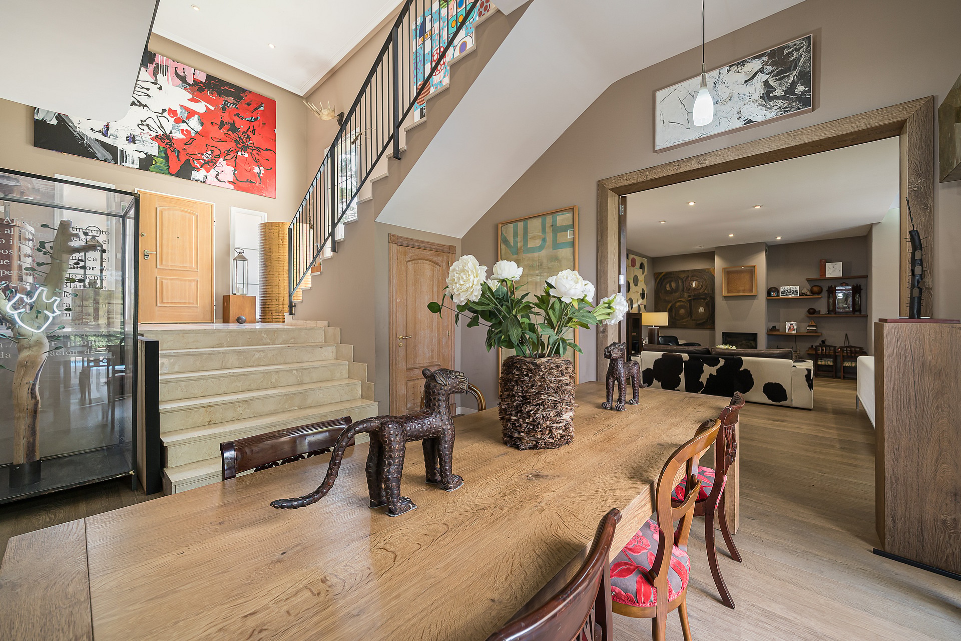Interior view of an entryway with a stone staircase, wooden dining table, and large floral arrangement, looking through to a living room.