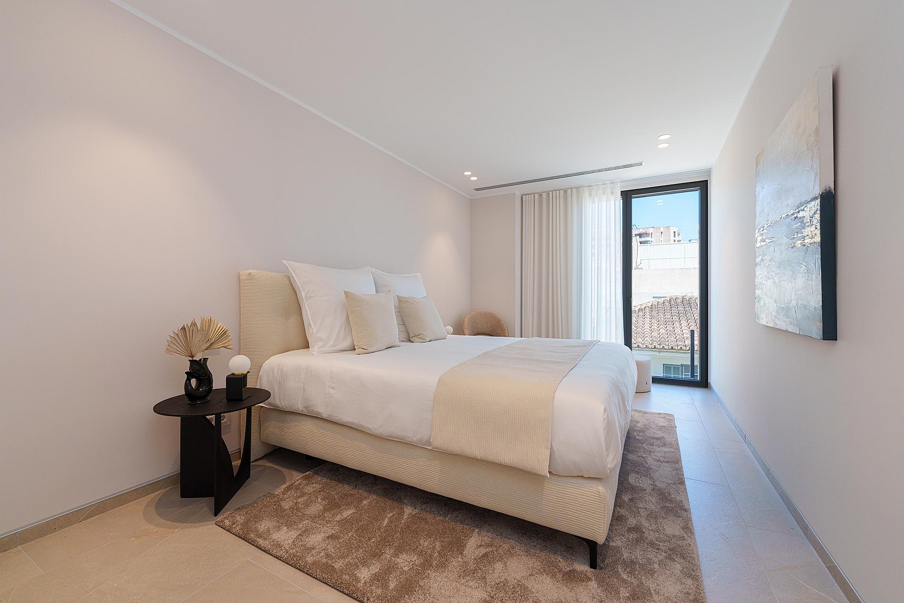 Dormitorio minimalista con cama en tonos neutros, mesita oscura y puerta de balcón con luz natural.