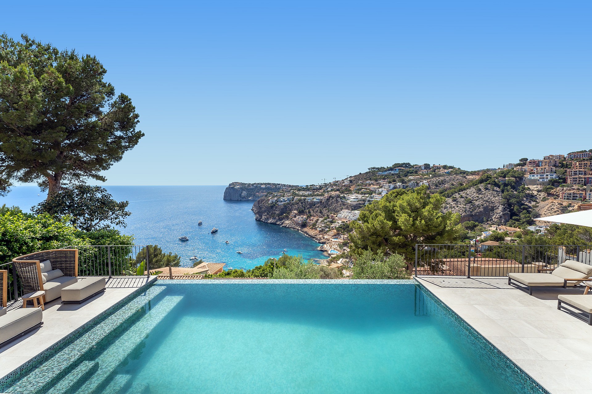 An infinity edge swimming pool with turquoise mosaic tiles overlooking a sea bay with rocky cliffs and pine trees.