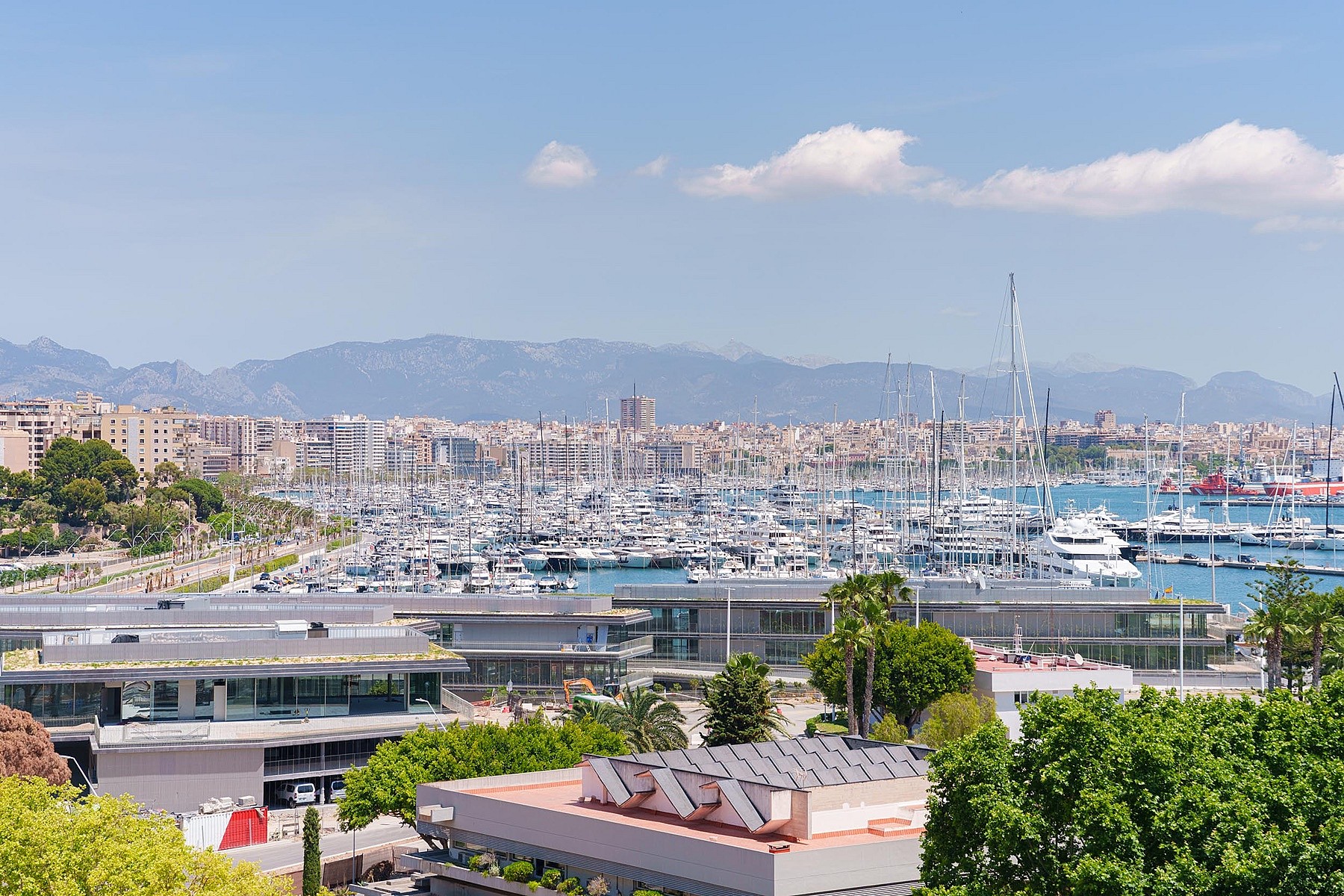 Vista elevada del puerto con yates, el perfil urbano y las montañas costeras bajo un cielo azul.