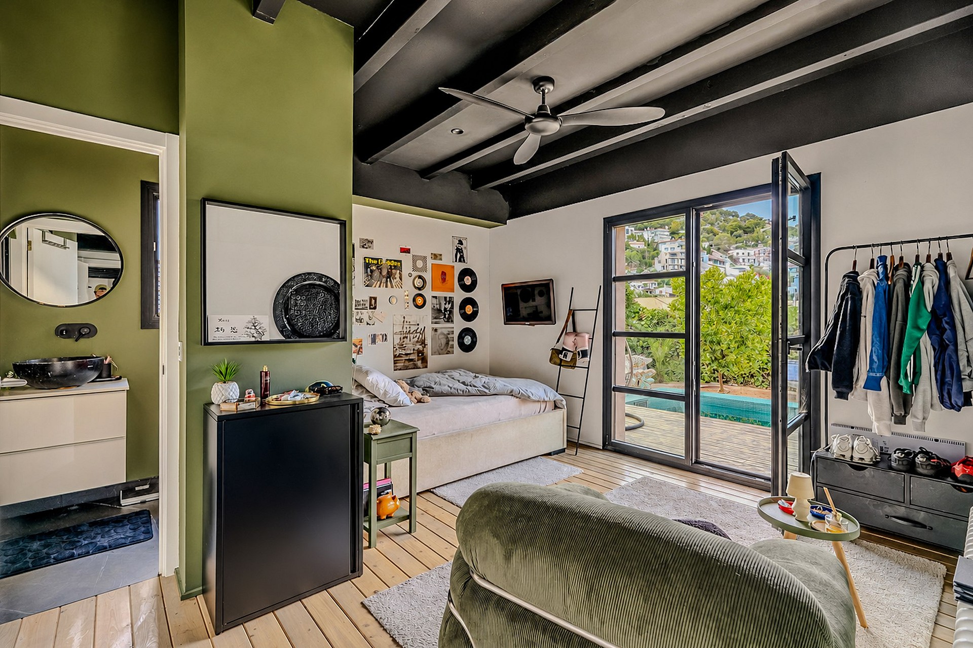 A bedroom with sage green walls, dark ceiling beams, a ceiling fan, and black framed glass doors opening to a garden patio.