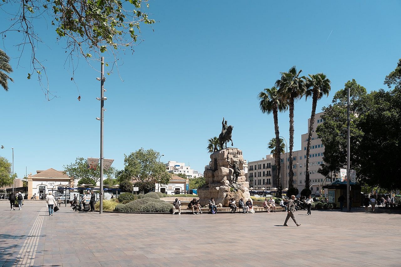 Wide shot of a sunny public square in Palma with palm trees, a central equestrian monument, and pedestrians.