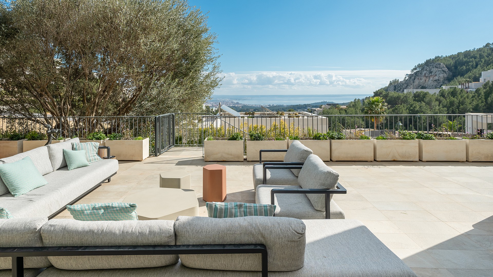 Outdoor terrace lounge with stone flooring, metal and fabric furniture, olive tree, and views of the Mediterranean coastline.