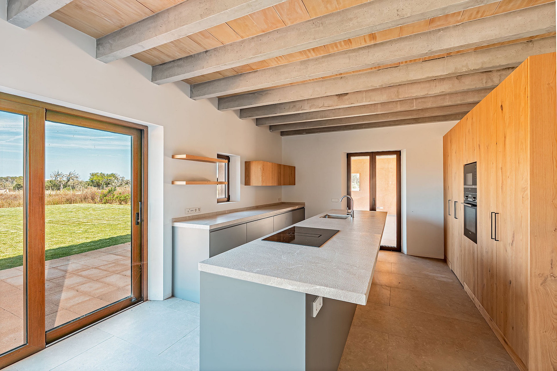 Modern kitchen featuring a central island, light grey cabinetry, wooden ceiling beams, and large glass doors overlooking a field.