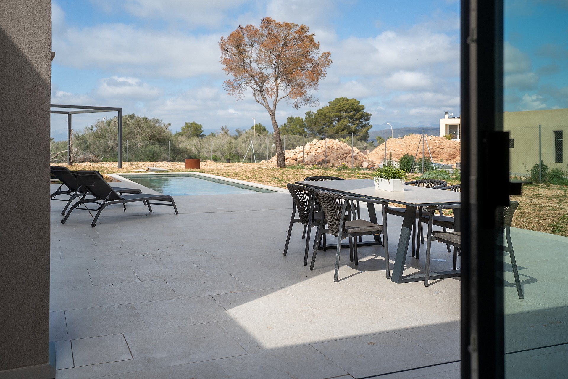 A covered terrace with a dining set and sun loungers overlooks a swimming pool and distant hills under a sunny sky.