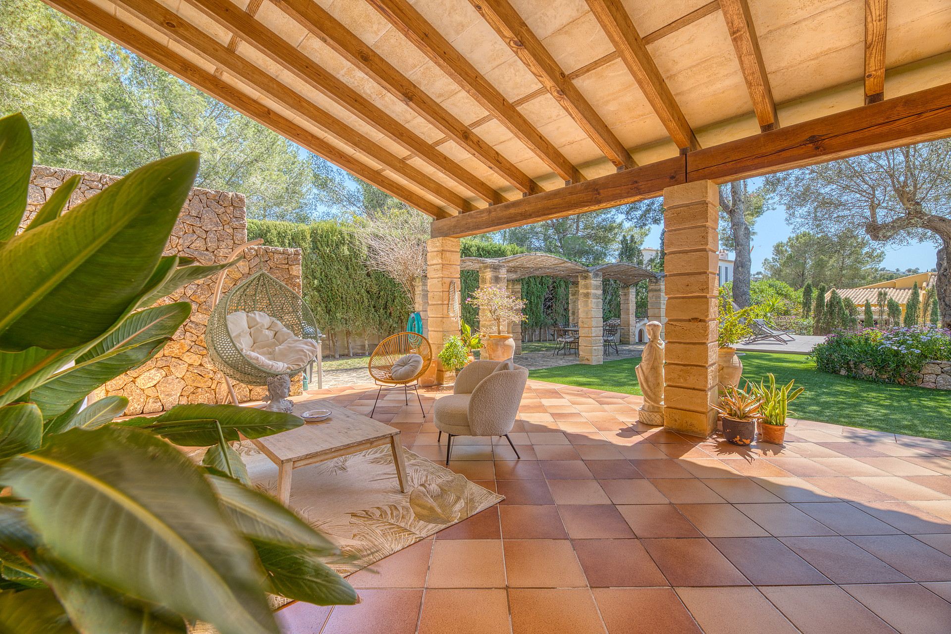 A covered terrace with terracotta floor tiles and wooden beam ceiling, featuring outdoor armchairs and a dry stone wall.