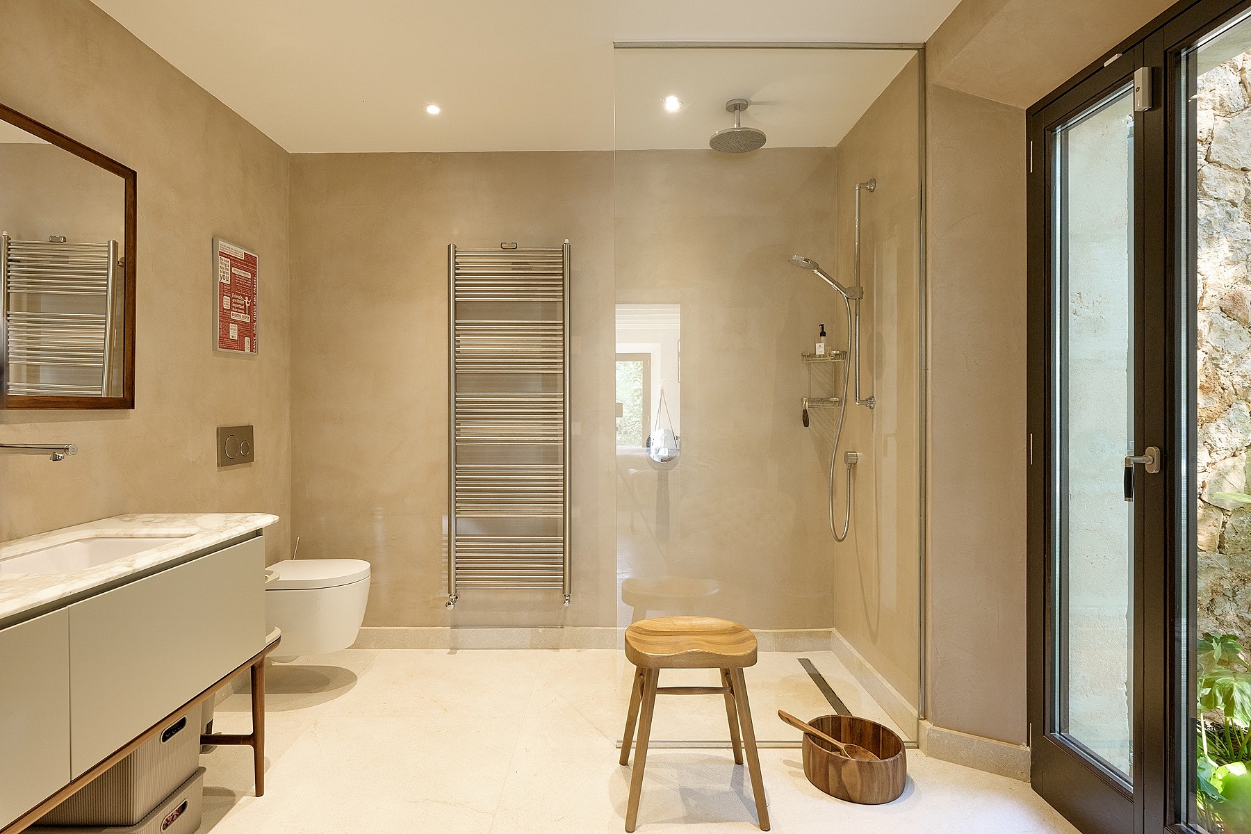 A minimalist bathroom with beige walls, a walk-in glass shower, wall-mounted vanity, and a wooden stool, featuring a glass door leading outside.