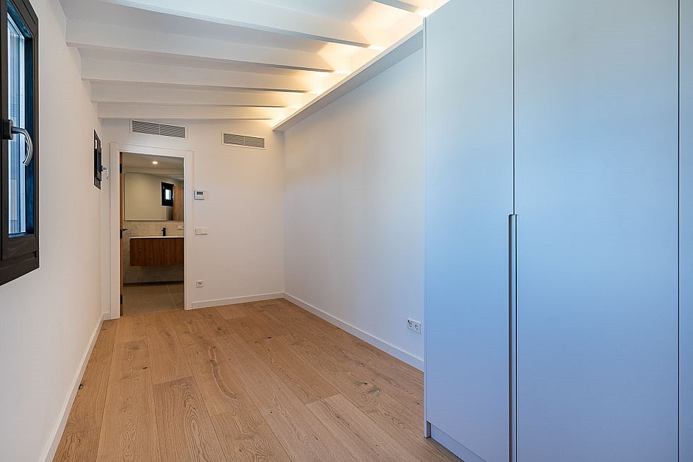 A minimalist bedroom interior featuring light wood flooring, white walls with beamed ceilings, and a tall integrated wardrobe.