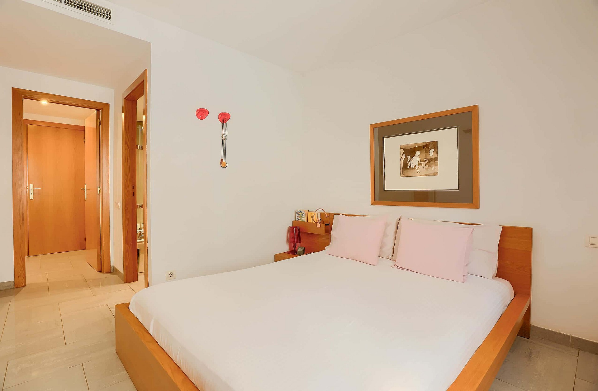 A double bedroom with a light wooden bed frame, white bedding, and pale pink pillows. Two wooden doors lead to adjacent rooms.