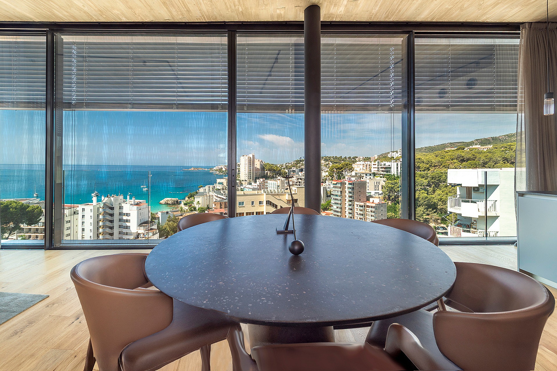 Round dark stone dining table surrounded by curved tan leather chairs in front of floor-to-ceiling windows overlooking the sea.