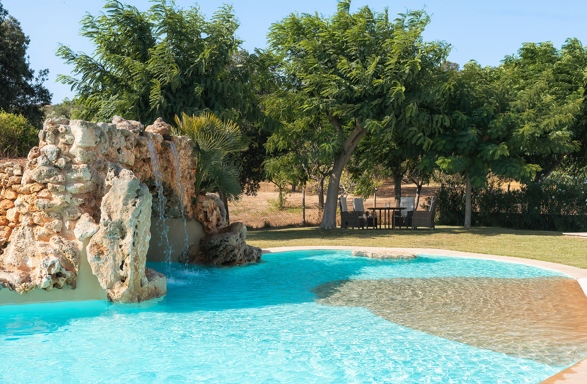 Piscina de superficie turquesa con cascada de roca natural, rodeada por árboles maduros.