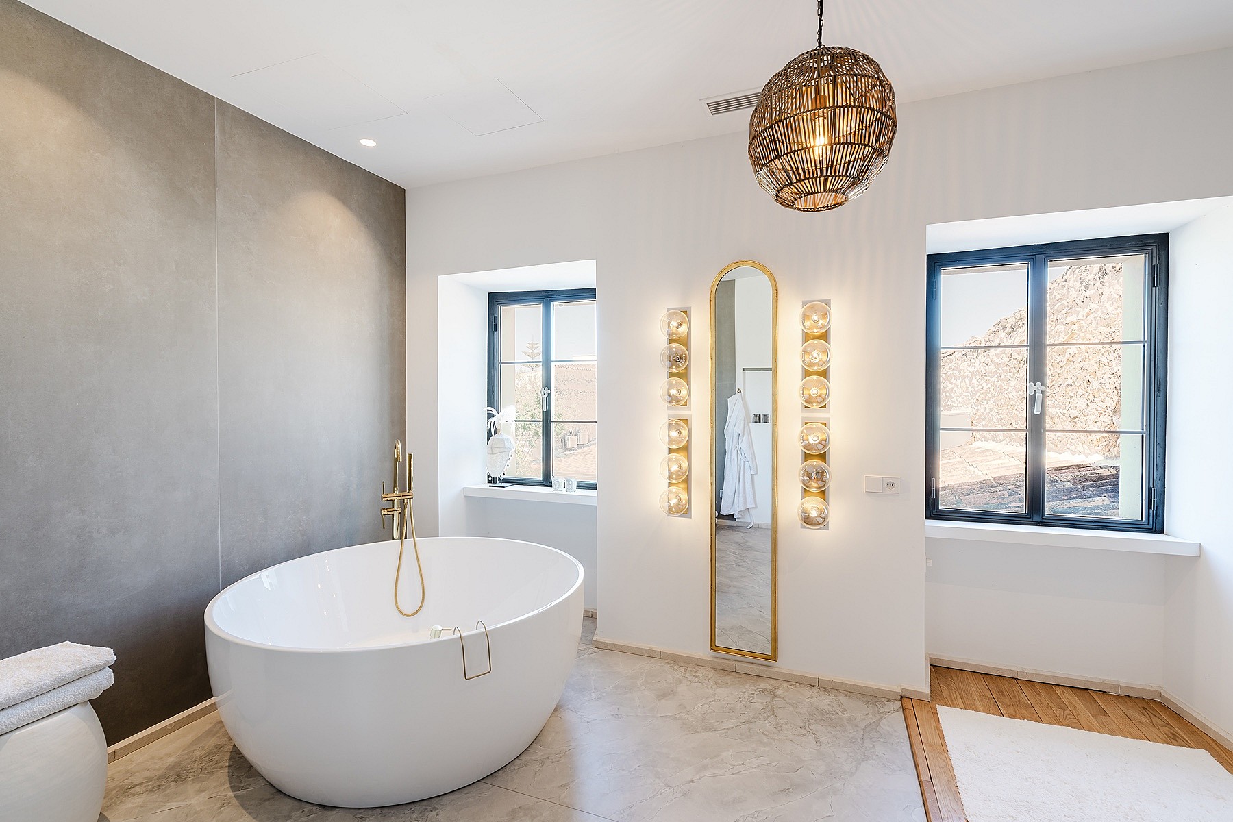 Modern bathroom featuring a white oval freestanding bathtub against a grey accent wall and two windows with mountain views.