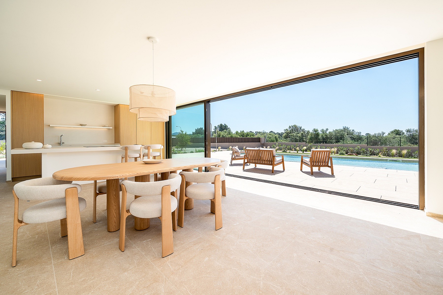 Dining area with oval light oak table and modern upholstered chairs opening onto a terrace with a swimming pool and trees.