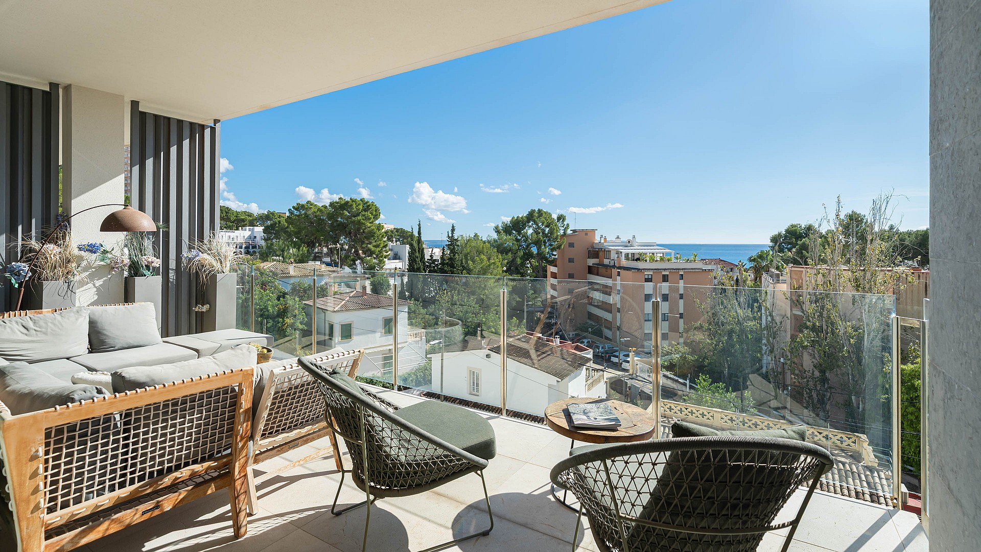 A covered terrace with grey outdoor furniture, glass railings, and a view of trees and houses under a clear blue sky.