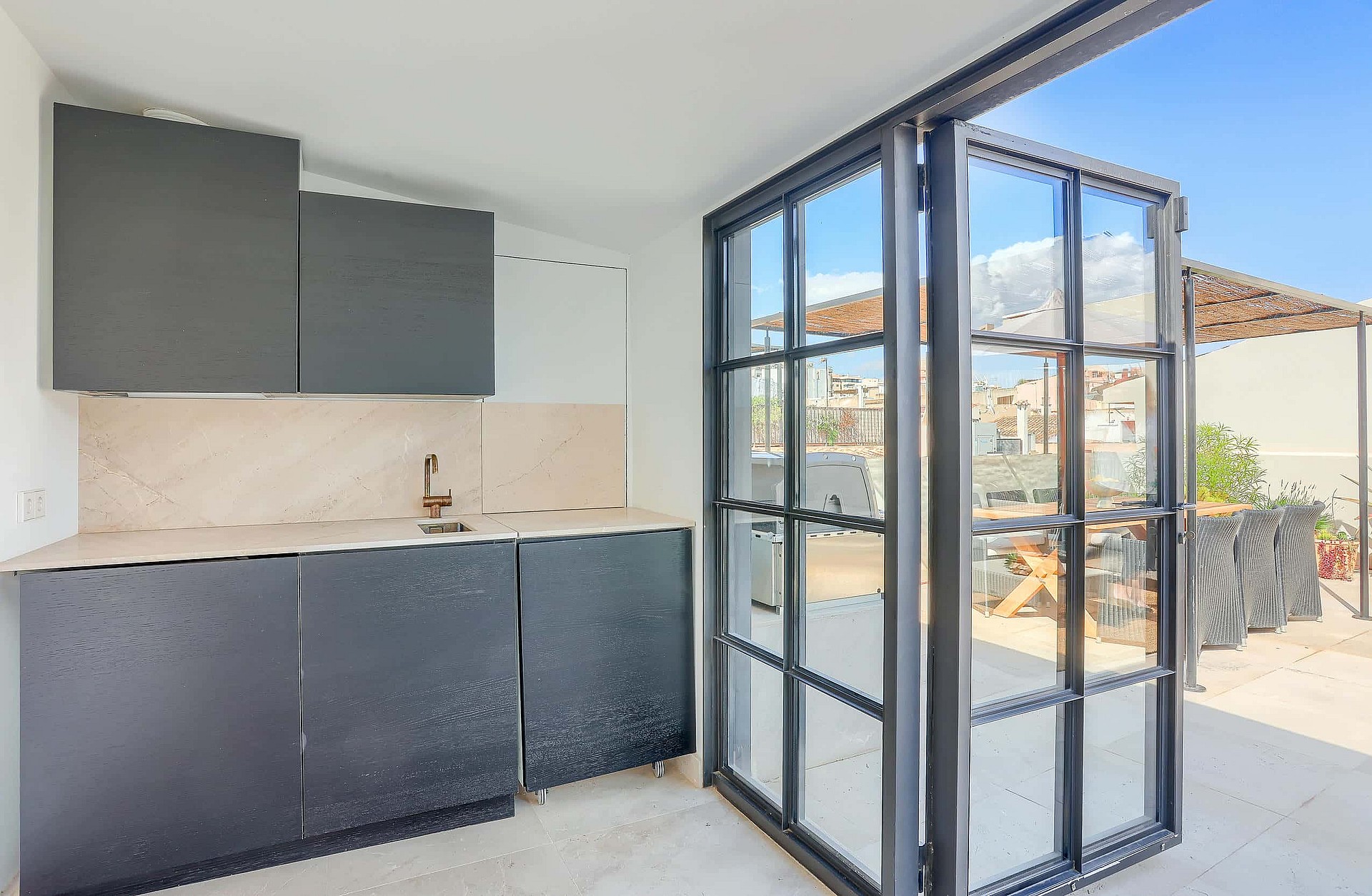 Compact outdoor kitchenette with charcoal cabinetry and stone backsplash adjacent to black-framed glass folding doors opening onto a terrace.