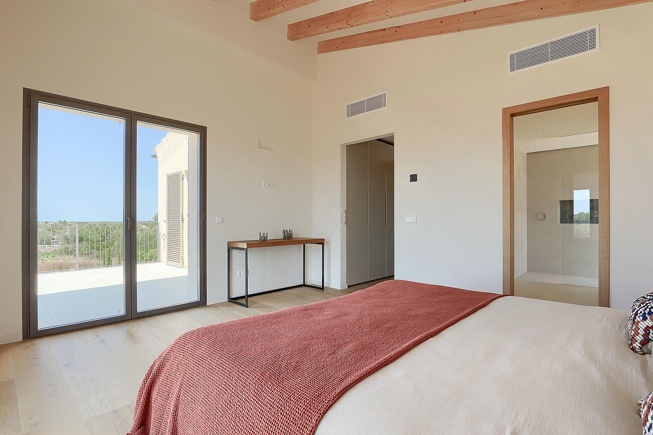 A bedroom with light wood floors, a rust-colored bed throw, and exposed timber ceiling beams, featuring sliding glass doors.