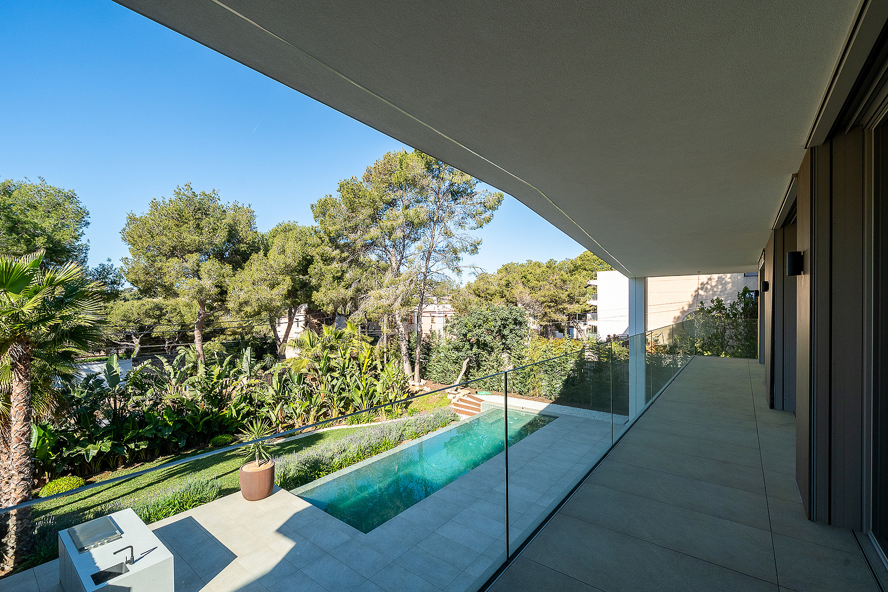 Covered concrete terrace with glass railing looking down onto a narrow rectangular lap pool surrounded by pine trees.
