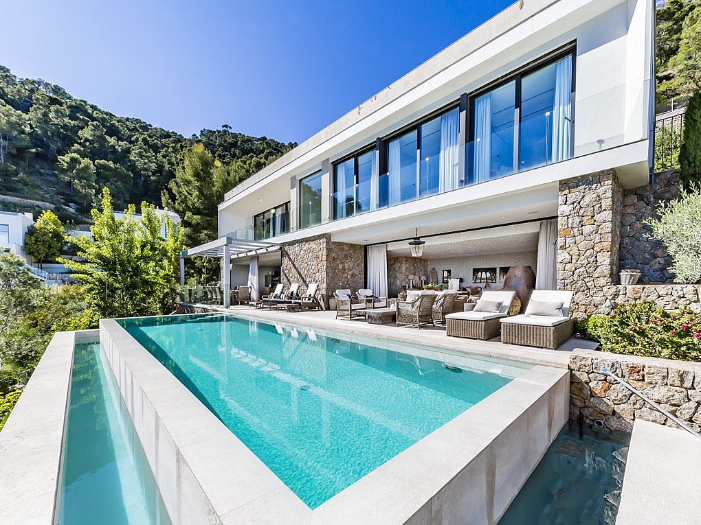 Exteriör av en modern vit villa med stenfasad, infinity-pool med turkost vatten och en möblerad terrass.