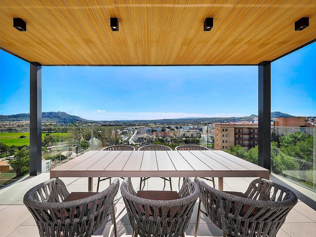 A covered terrace with a wood-paneled ceiling, a long dining table, and dark woven chairs overlooking a landscape of mountains.