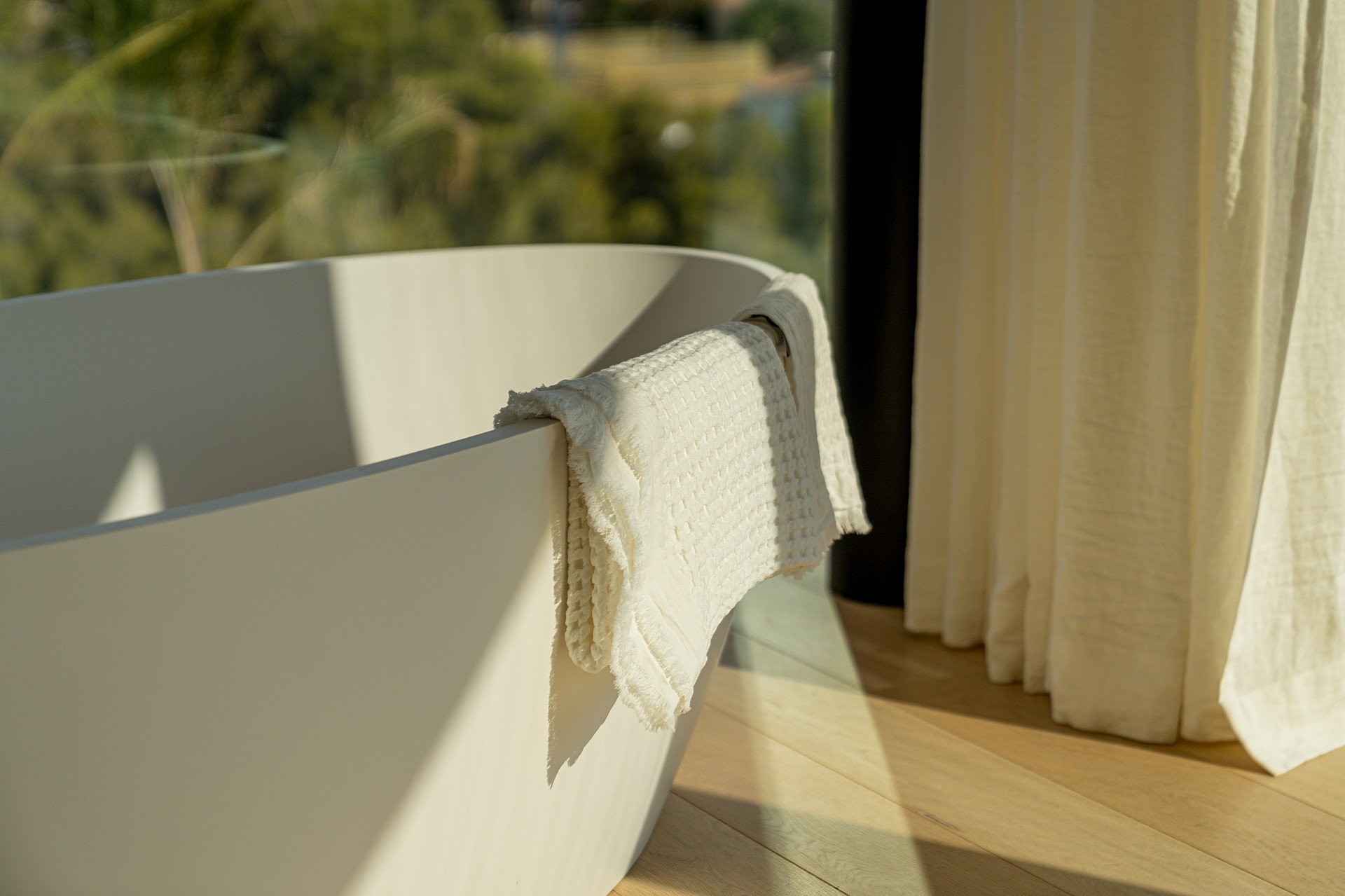 Close-up of a white freestanding bathtub with a textured waffle-weave towel draped over the rim, adjacent to floor-to-ceiling cream curtains and light oak flooring.
