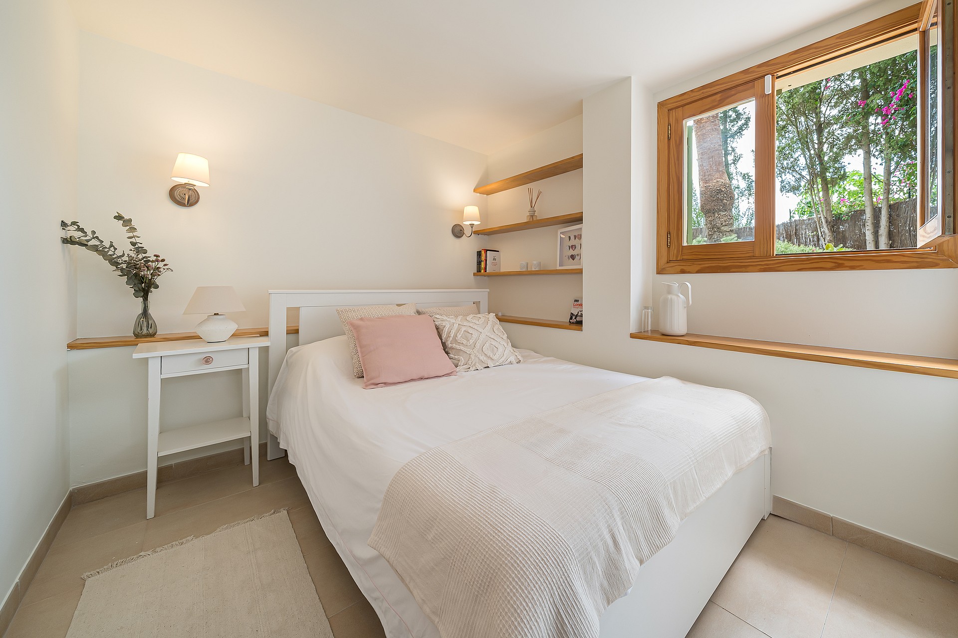 A bright bedroom featuring white linens, built-in wooden shelving, and a large timber-framed window facing garden trees.