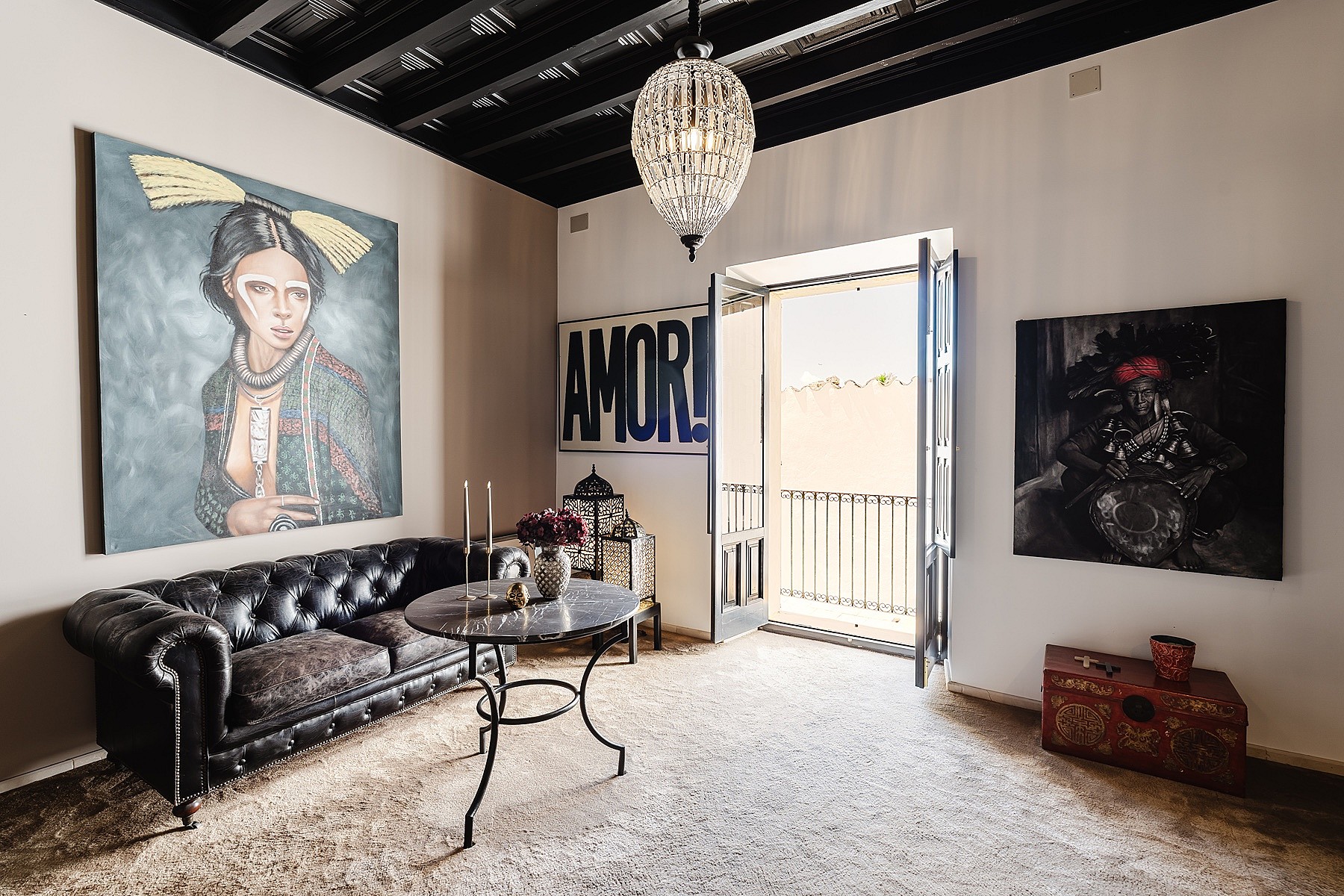 A living room in a Sencelles townhouse featuring a black leather tufted sofa, a glass-topped table, and large-scale artwork on white walls with a black exposed beam ceiling.