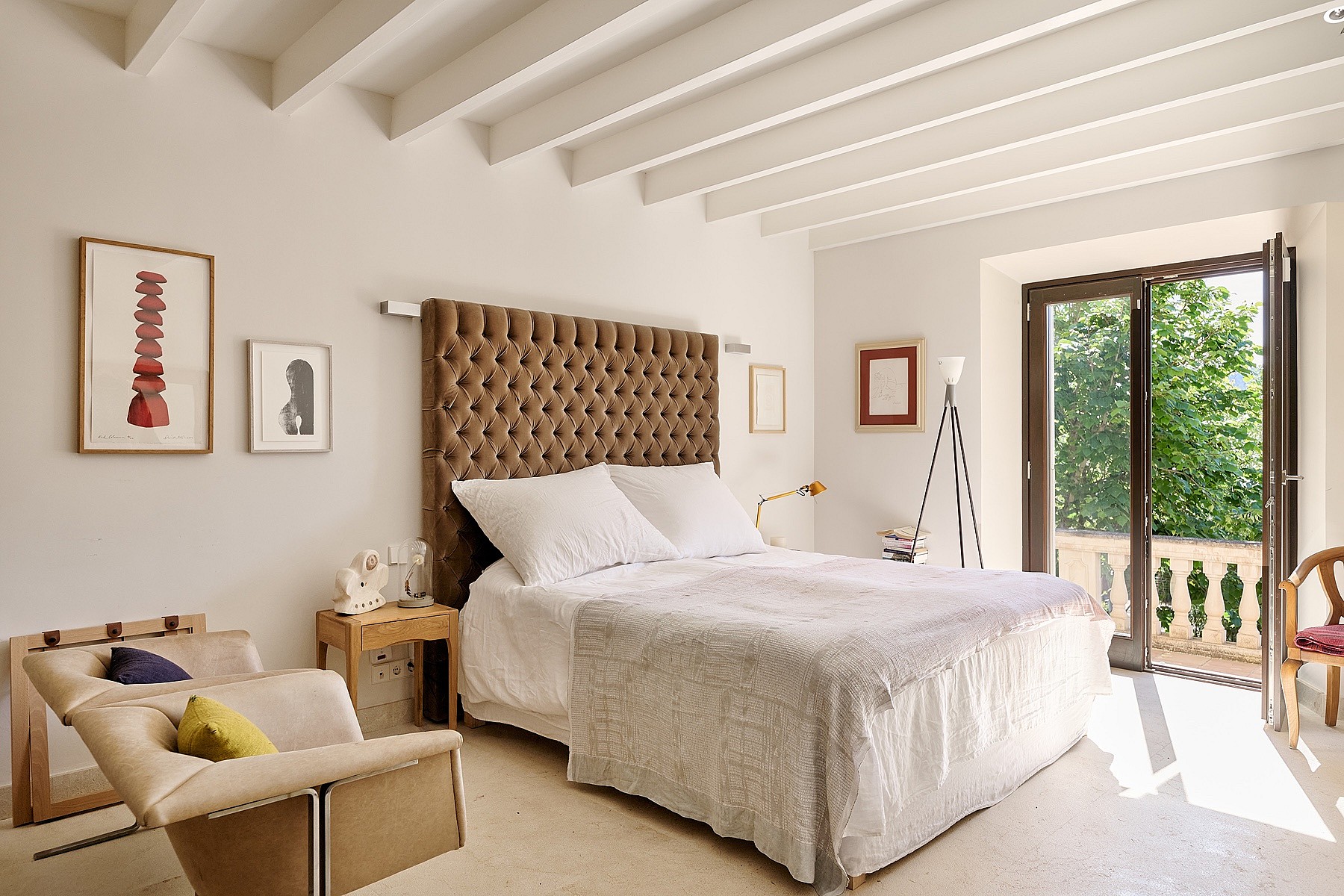 A bedroom with white walls, exposed wooden ceiling beams, a tufted brown headboard, and French doors leading to a terrace.