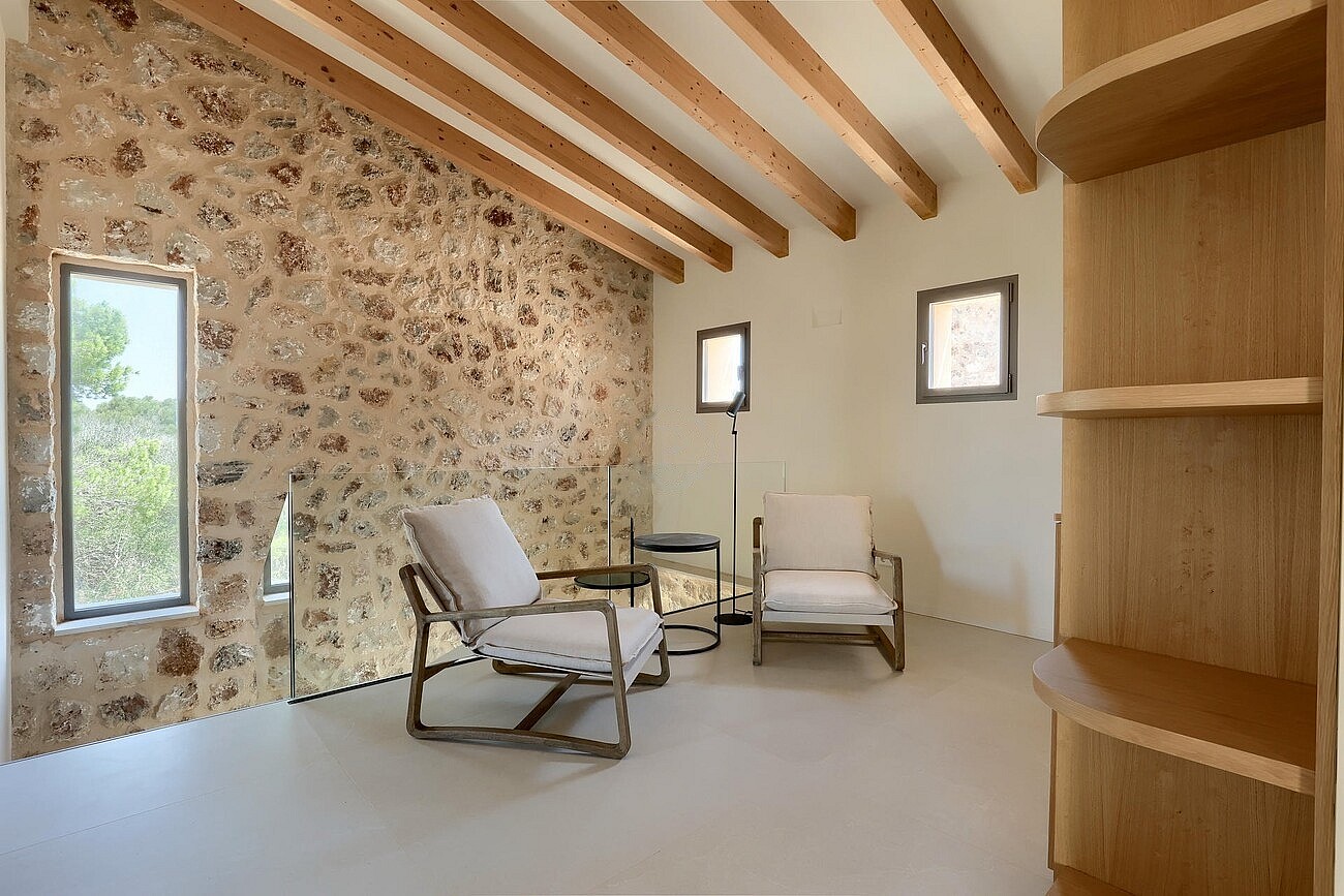 A living space featuring a textured dry-stone wall, exposed wooden ceiling beams, and two neutral-toned armchairs on a smooth floor.