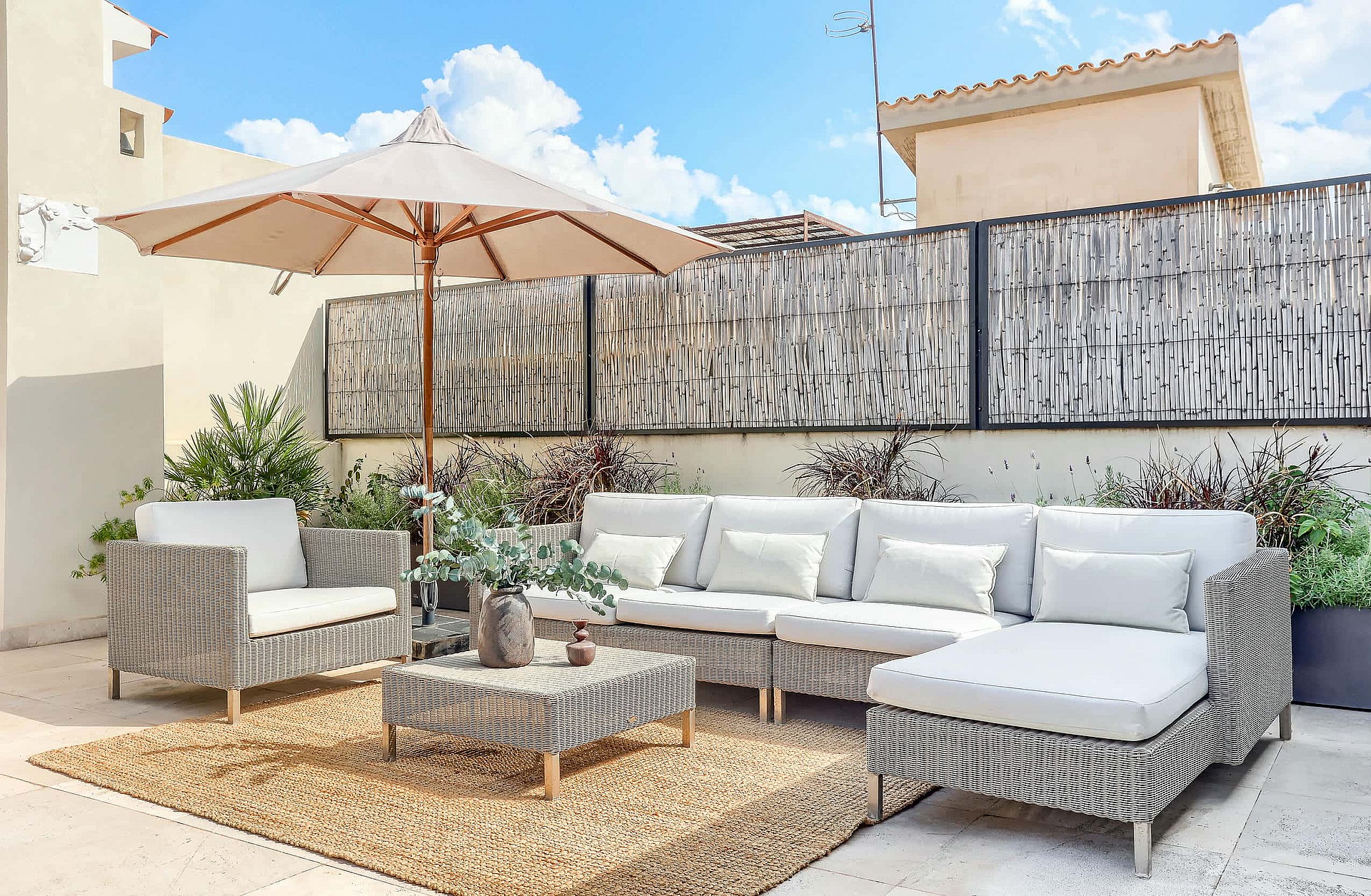 A rooftop terrace featuring a modular wicker sofa set with light cushions on a woven rug under a large beige umbrella.