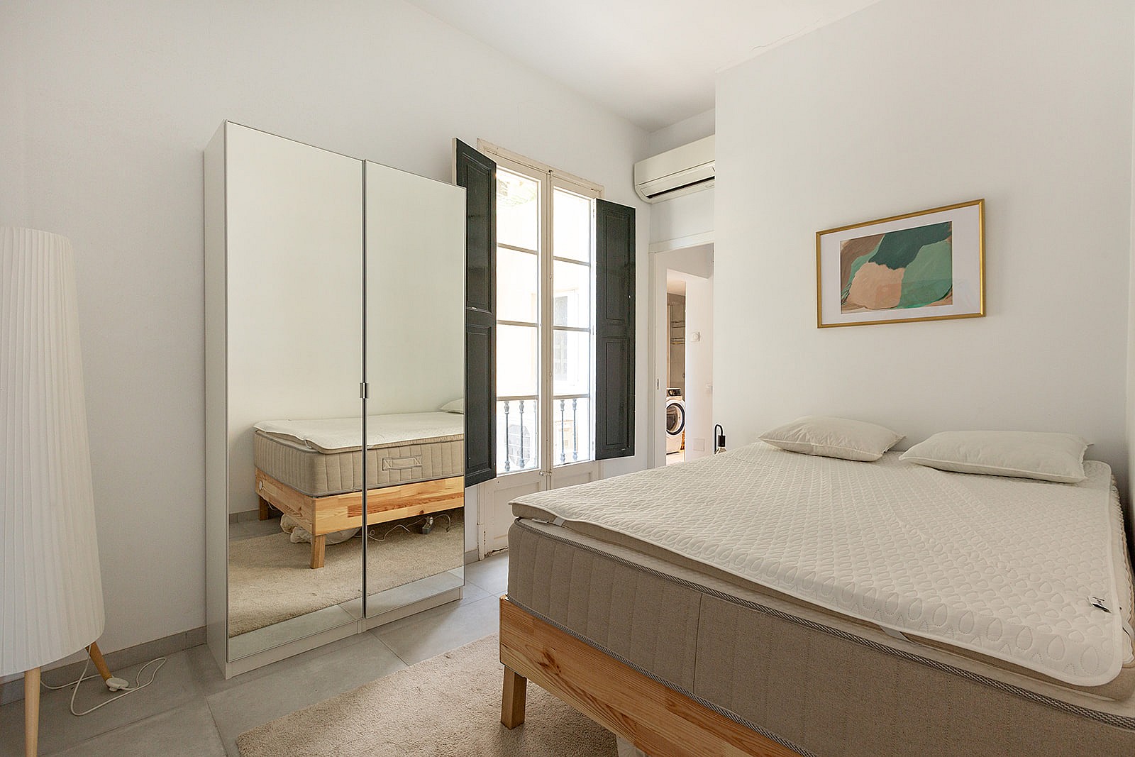 A bright bedroom with white walls, a double bed with a wooden frame, and a tall mirrored wardrobe by a window.