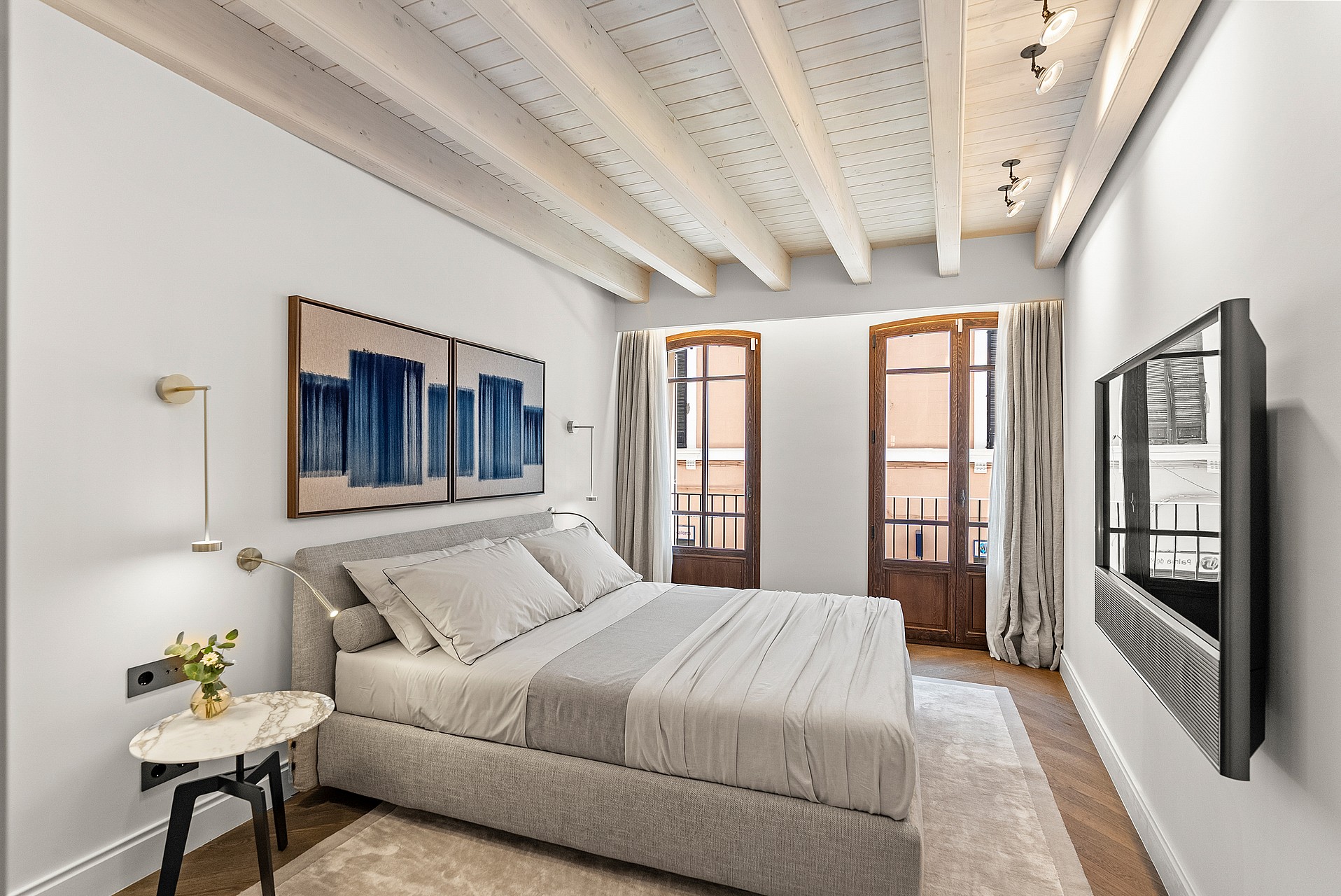 A bedroom featuring exposed timber ceiling beams, herringbone oak floors, and two arched glass doors opening to balconies.