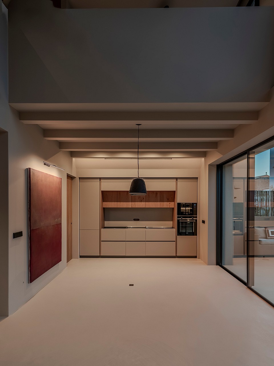 A modern kitchen featuring handleless beige cabinetry, exposed ceiling beams, and large sliding doors opening to a terrace.
